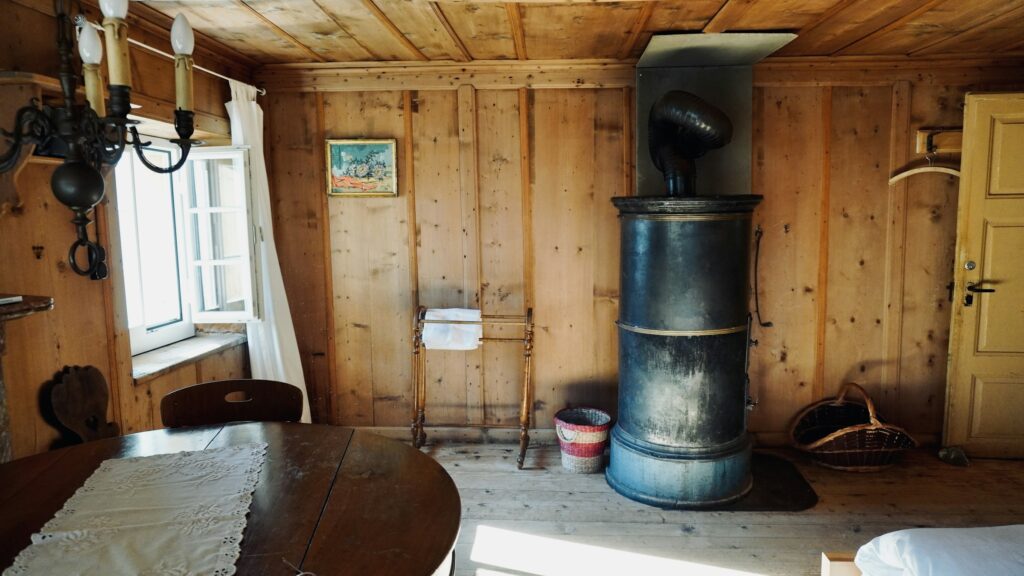 Hoher runder schwarzer Ofen in einer holzvertäfelten Stube, oben ein Rohrbogen in die Wand