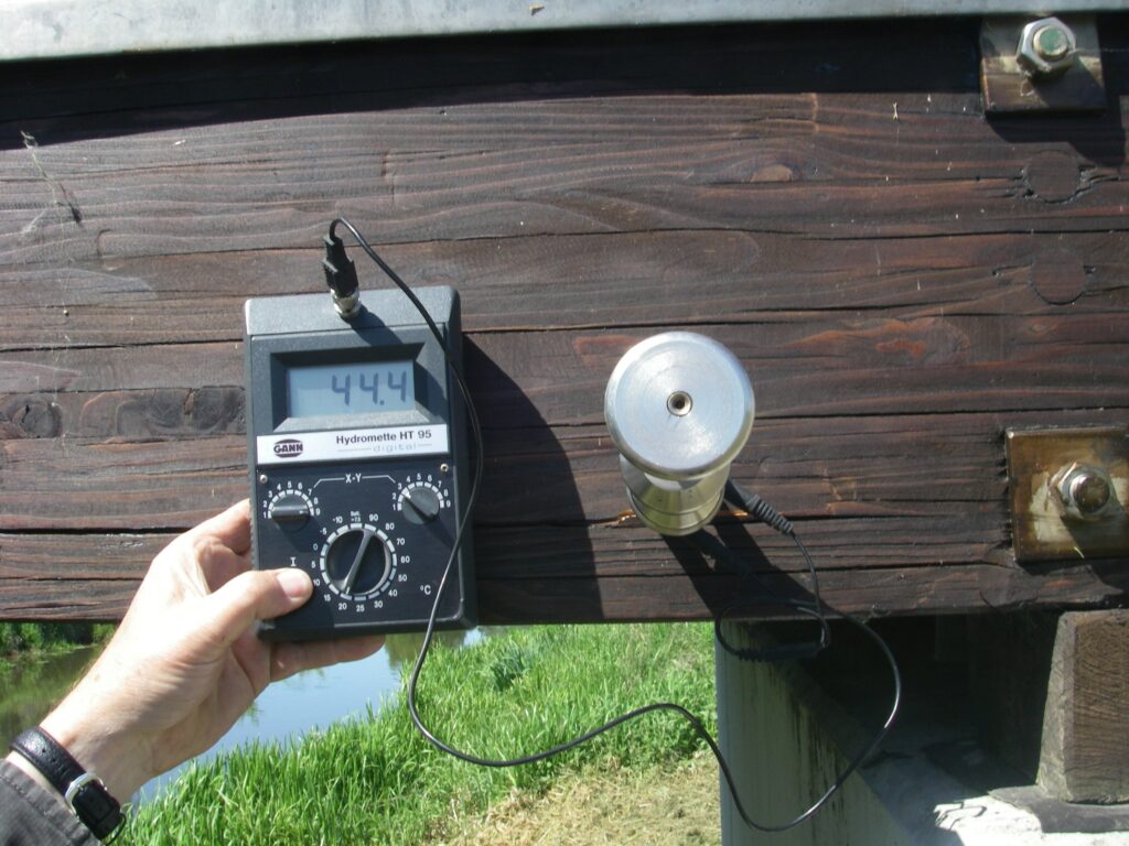 Hand hält ein Holzfeuchtemessgerät, dessen Elektroden in einem Holzbalken stecken